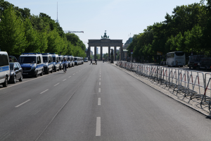 Eine Reihe von Polizeiwagen auf einer Straße vor dem Brandenburger Tor in Berlin geparkt, mit Menschen auf Fahrrädern und in der Nähe stehend, Barrieren, Bäumen und einem Bogen mit Statuen im Hintergrund.