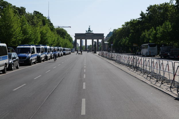 Eine Reihe von Polizeiwagen auf einer Straße vor dem Brandenburger Tor in Berlin geparkt, mit Menschen auf Fahrrädern und in der Nähe stehend, Barrieren, Bäumen und einem Bogen mit Statuen im Hintergrund.