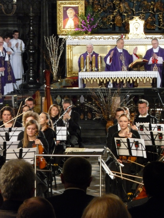 Eine Gruppe von Menschen, die Geigen spielen, mit Notenblättern vor sich, mit drei Kirchenvätern und einer Bibel im Hintergrund und einem Photo-Rahmen mit Verzierungen drumherum.