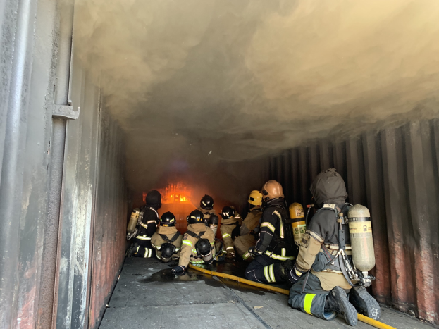Feuerwehrleute in Schutzausrüstung und Sauerstoffflaschen arbeiten in einem raucherfüllten Tunnel, mit einem sichtbaren Feuer im Hintergrund und einer Wand auf der linken Seite.
