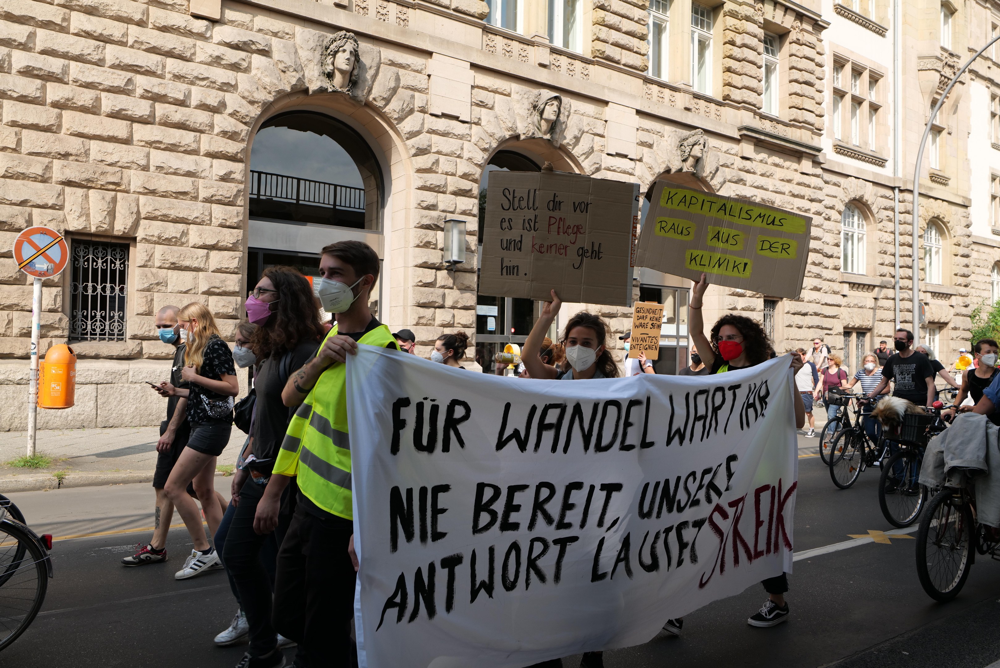 Eine Gruppe von Menschen marschiert bei einer Demonstration auf einer Stadtstraße, einige halten Schilder und andere fahren Fahrräder, im Hintergrund ein Gebäude mit Rundbogenfenstern, Säulen und Skulpturen.