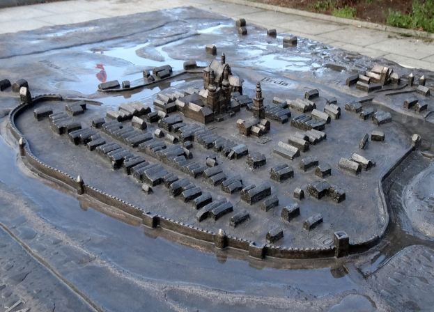 Eine Modellstadt in einer Pfütze umgeben von Gebäuden, Bäumen, Pflanzen und einer Wand.