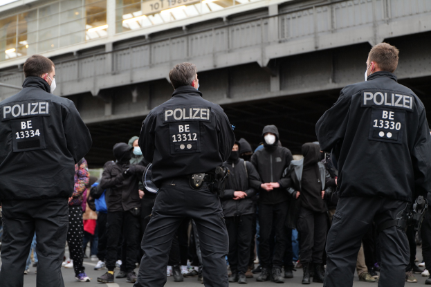 Polizisten in schwarzen Uniformen und Masken stehen vor einer Menge bei einer Demonstration, mit einer Brücke und einem Gebäude im Hintergrund.