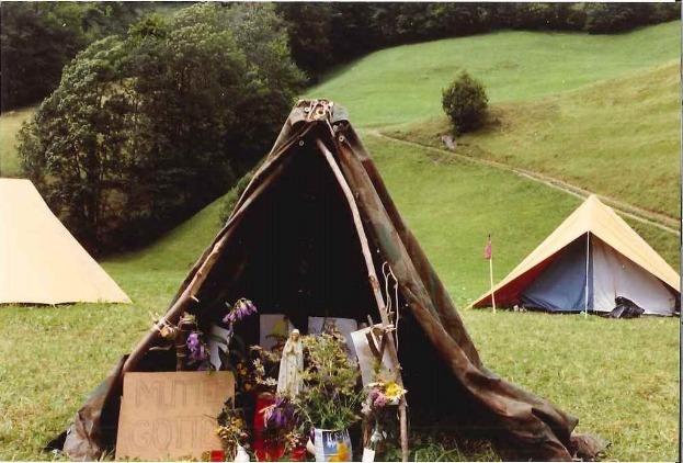 Mehrere Zelte und Blumenkübel sind auf Gras aufgestellt, mit einer Tafel im Vordergrund und Bäumen im Hintergrund.