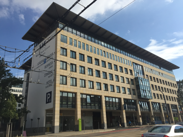 Das Stadthallen-Zentrum für Geschäftsleute in Köln, Deutschland, ein großes Gebäude mit vielen Fenstern, umgeben von Bäumen, Straßenlaternen, Strommasten, Stromkabeln, Kraftfahrzeugen auf der Straße und Menschen auf dem Gehweg, mit einem bewölkten Himmel im Hintergrund.