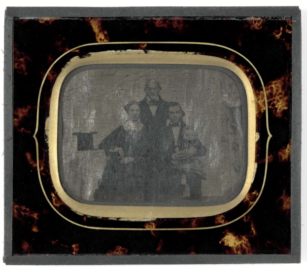 Altes Schwarz-Weiß-Foto eines formell gekleideten Mannes in Anzug und Krawatte und einer Frau in unbekannter Kleidung nebeneinanderstehend mit ernsten Gesichtern vor einem Fernseher vor einem weißen Hintergrund.