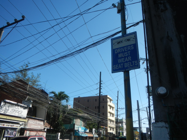 Eine Stadtstraße mit fahrenden Autos, Strommasten mit Drähten, Gebäuden mit Fenstern, Bäumen, Namensschildern und einem Himmel als Hintergrund, mit einem "Fahrer müssen den Sicherheitsgurt anlegen"-Schild an einem Strommasten.