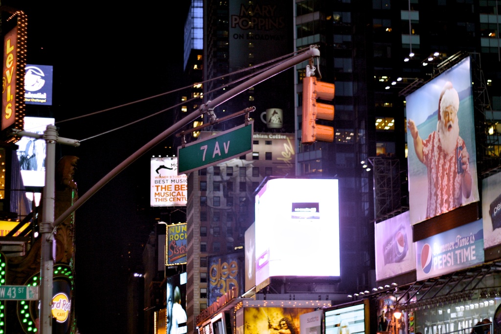 Eine nächtliche Stadtansicht mit Gebäuden und Werbetafeln auf beiden Seiten, einem Pfahl und einer Ampel auf der linken Seite, einem grünen Schild mit Text und einem weißen Schild mit Text in der Mitte.