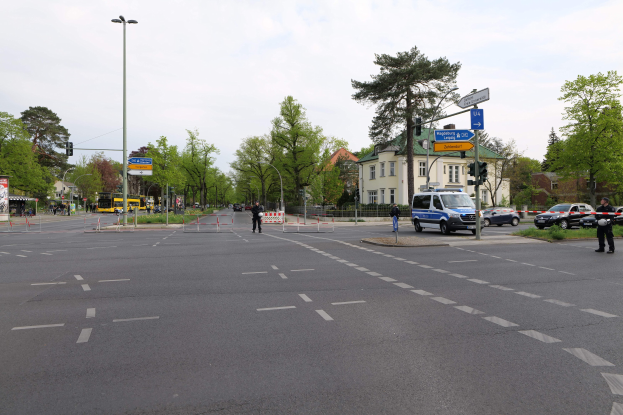 Ein Kreisverkehr mit einem parkenden Polizeiwagen, fahrenden Fahrzeugen, Fußgängern auf dem Gehweg, Verkehrsampeln, Bäumen, Gebäuden und einem bewölkten Himmel.