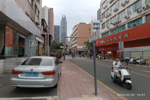 Ein Mann fährt mit einem Scooter eine Straße mit hohen Gebäuden entlang, mit Menschen auf den Bürgersteigen und Fahrzeugen auf der Straße, unter einem klaren blauen Himmel mit Bäumen im Hintergrund.