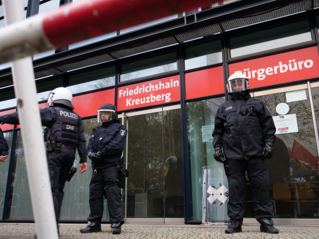 Polizisten in Helmen und Handschuhen vor einem Glasgebäude mit angeklebten Papieren und einem Pfahl links, während Schilder im Hintergrund "Friedrichshain Kreuzberg" anzeigen.