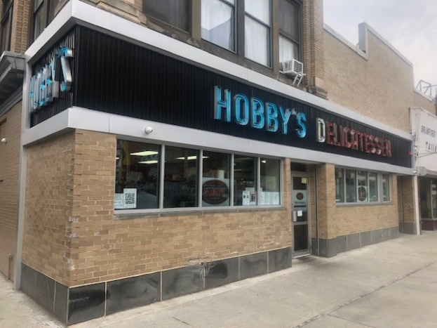 Außensicht von Hobby's Delicatessen in Chicago, Illinois, mit Glasfenstern und -türen, einem Namensschild, einem Laternenmast davor und einem bewölkten Himmel im Hintergrund.