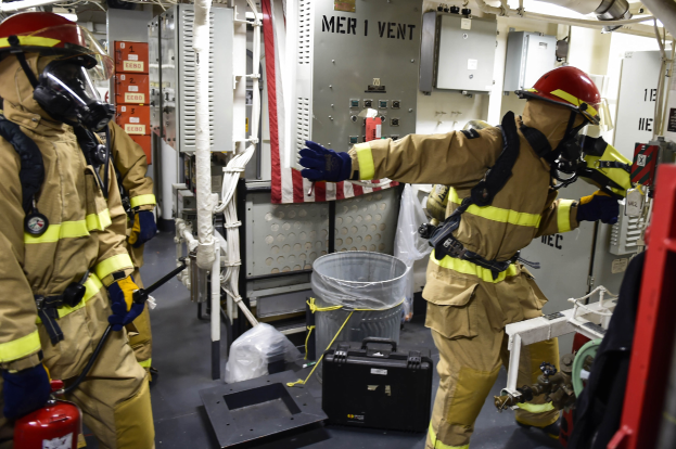 Drei Feuerwehrleute in Schutzausrüstung stehen zusammen in einem Raum mit einem Feuerlöscher links, einer Maschine rechts und diversen Gegenständen und Rohren im Hintergrund.