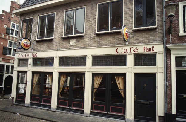 Ein Gebäude mit Fenstern und Türen, ein Schild mit der Aufschrift "Cafe Plan C in Amsterdam, Niederlande", ein Straßenschild, Bäume und ein bewölkter Himmel.