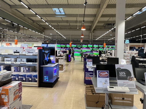 Ein großer Raum in einem Walmart-Geschäft, der mit Regalen voller Elektronik, darunter Computer und Drucker, gefüllt ist, mit Menschen auf dem Boden, einer Säule, Schildern, Deckenleuchten und Deckenventilatoren.