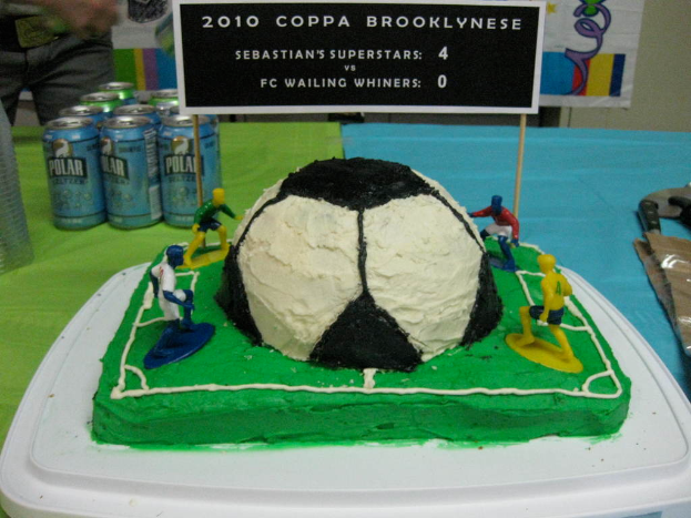Ein fußballstadion-themenkuchen mit einem fußball und vier spielern, einer banner, soft drink dosen und einer person in der oberen linken ecke.
