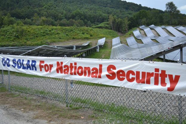 Ein Zaun mit einer Fahne, auf der "Geht Solar für die nationale Sicherheit" steht, vor einem Solarpanel-Feld, Bäumen, Pflanzen und Gras unter einem klaren blauen Himmel.