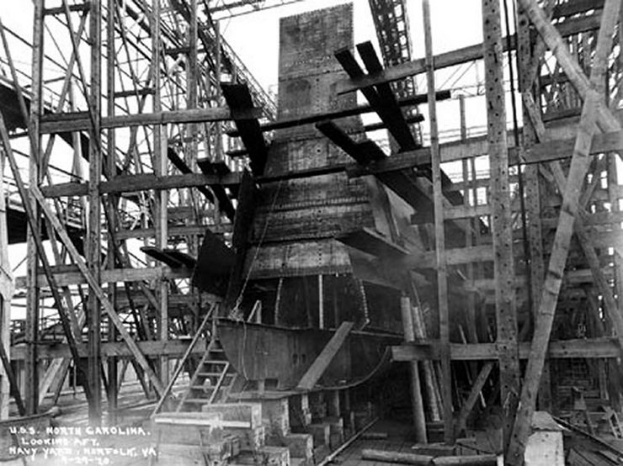 Schwarzes und weißes Foto eines Schiffes in Bau in einer Werft, mit hölzernen Pfählen und Treppen im Vordergrund und Himmel im Hintergrund, mit Text "Naval Weaponry" in der linken unteren Ecke.