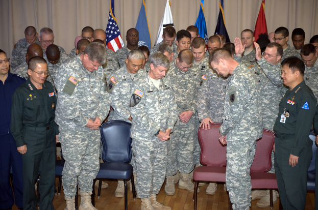 Eine Gruppe von Menschen, mostly in military dress, stand between red and blue chairs with flags in the background.