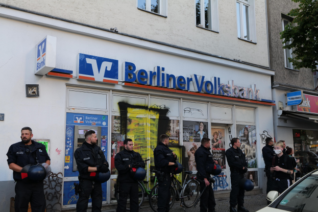 Gruppe von Polizisten vor einer Berliner Volksbank stehend, einige mit Helmen, mit einem Auto rechts und einem Baum links, Gebäude mit Fenstern und Texttafeln im Hintergrund.