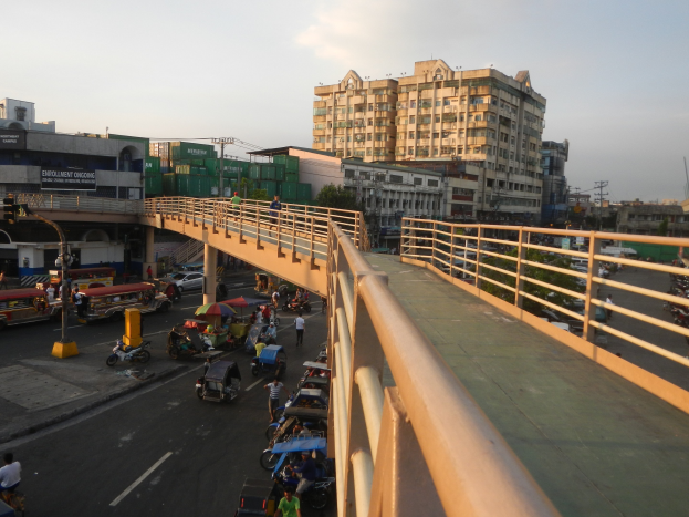 Vielbefahrene Straßenszene mit Brücke darüber, Fahrzeugen, Fußgängern mit Schirmen, Geländern, Gebäuden, Bäumen, Strommasten und einem klaren blauen Himmel.