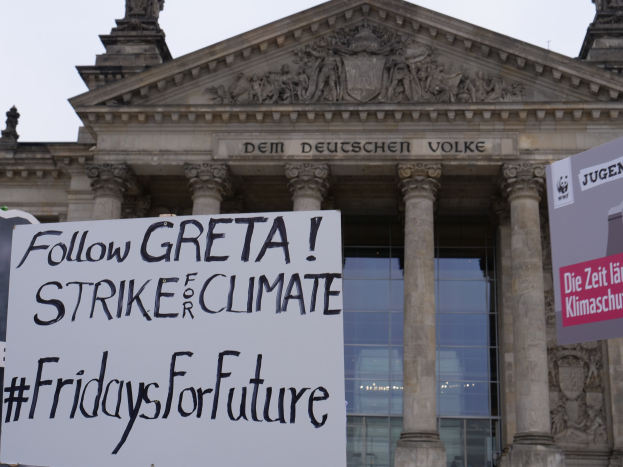 Großes Gebäude mit klassischer Architektur, das Säulen und Skulpturen aufweist, mit einem Protestschild, auf dem "Follow Greta Strike for Climate Fridays for Future" steht, vor einem klaren blauen Himmel.