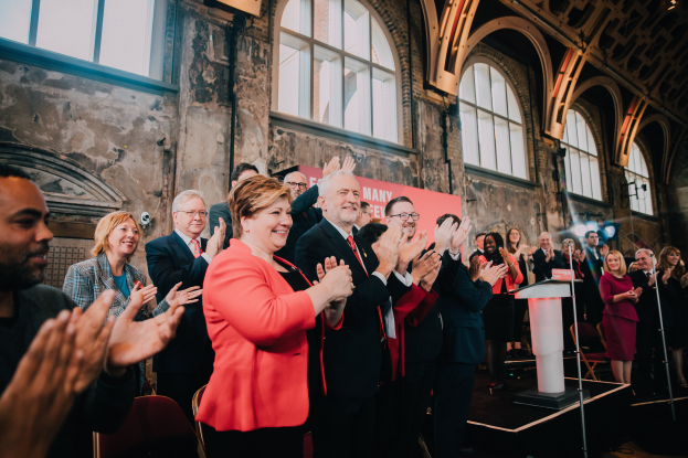 Eine Gruppe von Menschen steht vor einer Menge, die Hände klappend, mit einem Podium mit Mikrofon auf der rechten Seite und einem Banner im Hintergrund, das 'Covid-19 New York City Mayor Bill de Blasio' lautet.