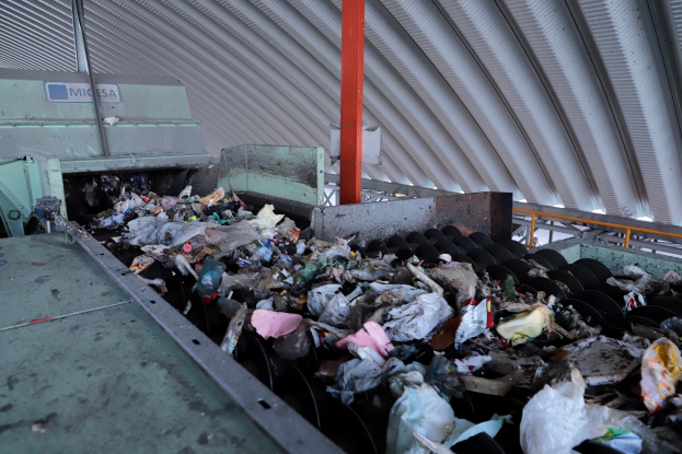 Ein großer Haufen Müll auf einem Förderband in einer Recyclinganlage, mit Maschinen im Vordergrund und einem Metalldach darüber.