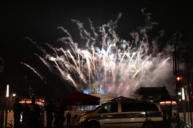 Ein Polizeiwagen steht vor einer Menschenmenge, mit beleuchteten Gebäuden, Bäumen, Pfählen, Verkehrszeichen und Schildern im Hintergrund, während farbige Feuerwerke den Himmel erhellen während einer Silvesterfeier in Berlin.