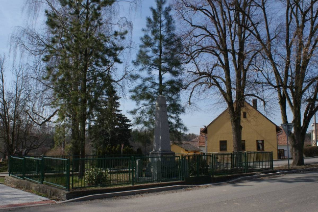 Eine Straßenszene mit einem Holocaust-Gedenkmonument in der Mitte, umgeben von einem Metallzaun, Pflanzen, Gras, Bäumen, Häusern, einem Straßenpfahl und einem bewölkten Himmel.