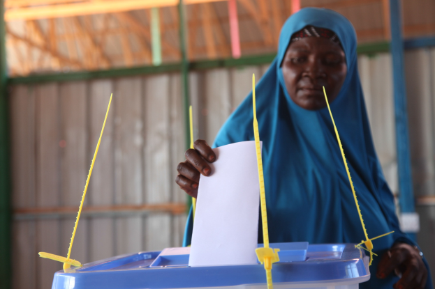 Frau mit blauem Kopftuch, die einen Zettel an einer Wahllokal-Urne hält.