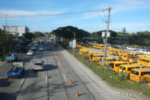 Eine belebte Straßenszene mit verschiedenen Fahrzeugen, Verkehrskegeln, Laternenmasten, Strommasten mit Kabeln und Texttafeln, mit Maschendrahtzaun rechts, Bäumen, Gebäuden und einem bewölkten Himmel im Hintergrund.