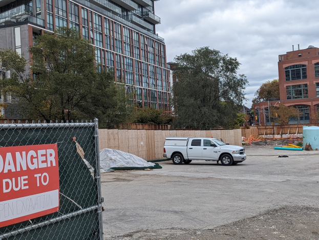 Truck parked in front of a building with a "Danger Due to Open Excavation" sign, trees and buildings in the background under a clear blue sky.