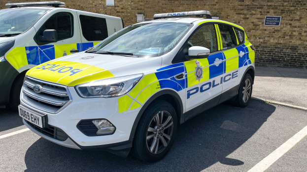 Zwei Polizeiautos nebeneinander auf einem Parkplatz geparkt, mit einer Wand und einer Texttafel im Hintergrund.