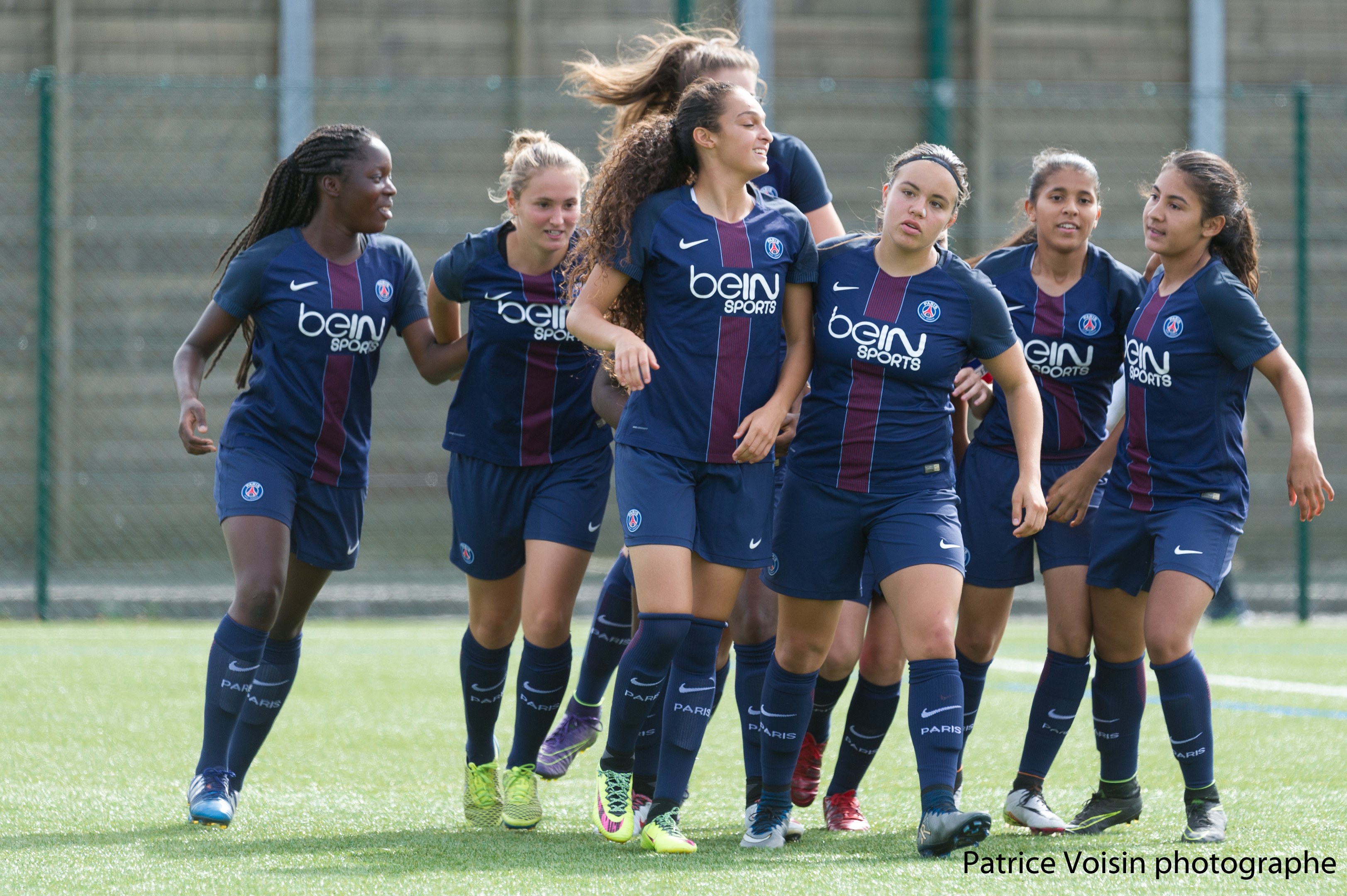 Gruppe junger Frauen beim Fußballspielen auf einem Rasenplatz mit Maschendrahtzaun und einer Wand im Hintergrund, Text in der rechten unteren Ecke lautet "Paris Saint-Germain Frauenfußball".