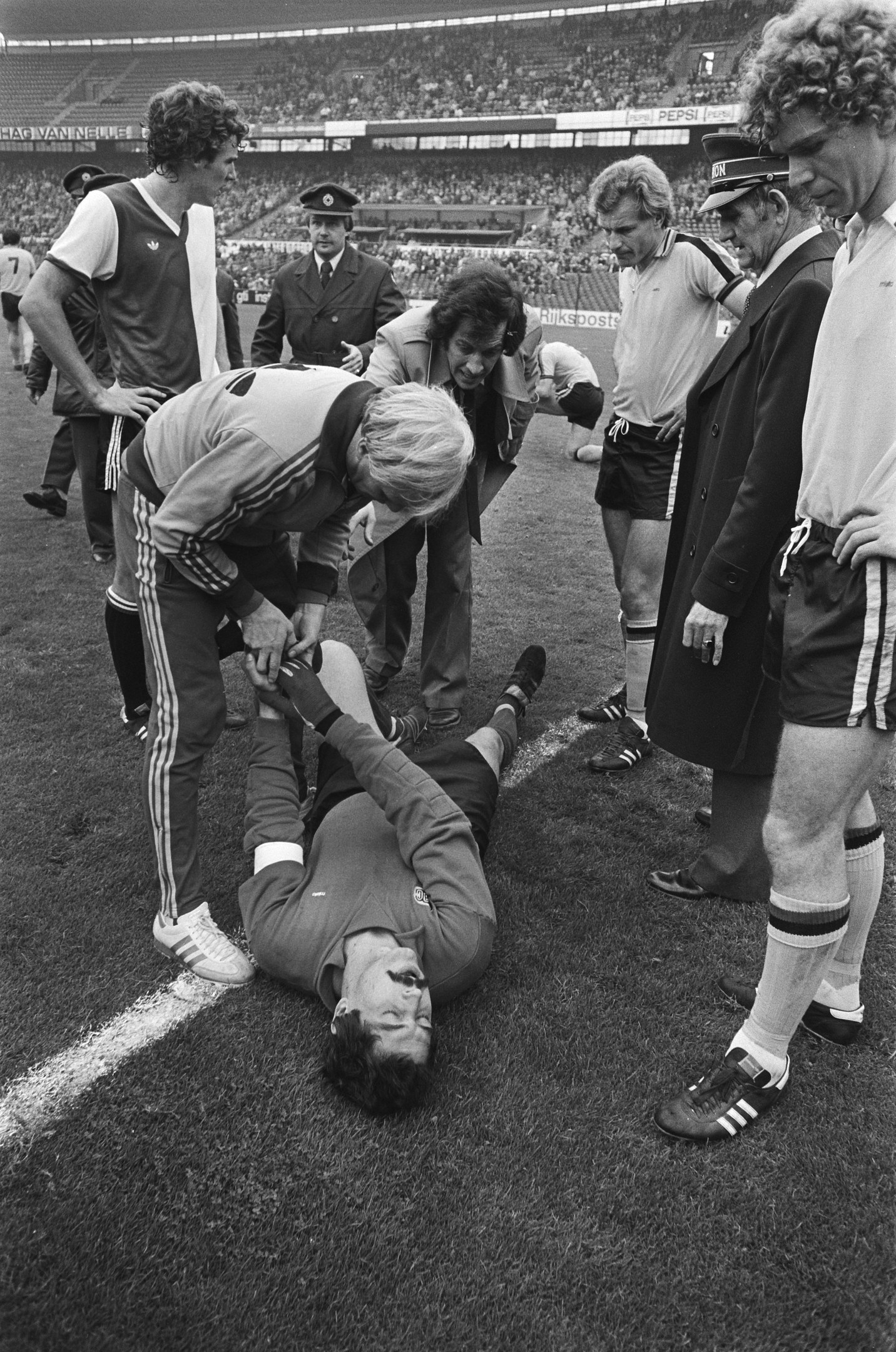Schwarzes und weißes Foto von Fussballspielern und Stabmitarbeitern, die sich um einen verletzten Spieler auf dem Feld versammeln, mit einer Zuschauermenge im Hintergrund.